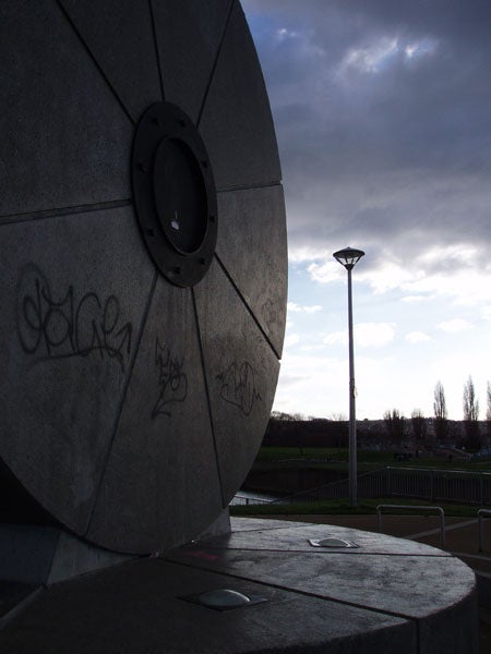 Photograph taken with an Olympus µ (Mju) Digital 500 camera showcasing a concrete structure with graffiti and a solitary street lamp against a cloudy sky.