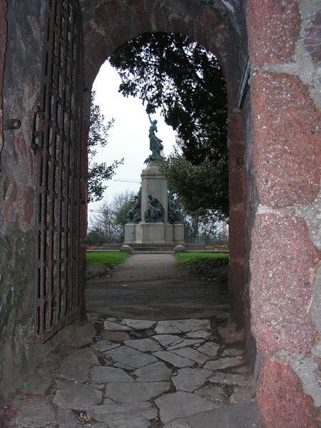 Photo taken with the Konica Minolta DiMAGE Z20 camera showing a view through an old arched gateway with an open iron gate, leading to a statue in the distance. The image showcases the camera's ability to capture the texture of the stone pathway and the contrast between the shaded archway and the lit background.