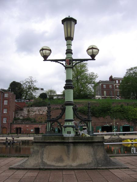 Photograph taken with a Canon PowerShot A510 digital camera featuring a vintage street lamp in the foreground with a serene riverside and buildings in the background under a cloudy sky.