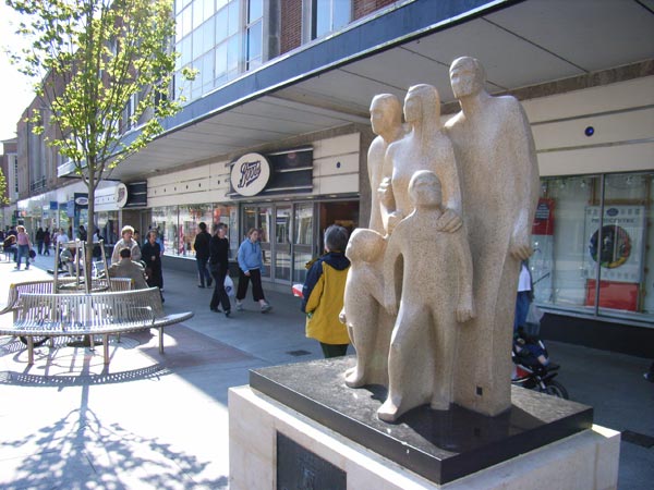 Photograph taken with a Ricoh Caplio R1V Digital Camera featuring a stone sculpture of a family in an urban pedestrian area, with shoppers walking by and a clear blue sky above.