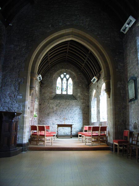Interior of a stone-walled church with arches, taken with a Ricoh Caplio R1V digital camera, showcasing chairs arranged near an altar with stained glass windows in the background.