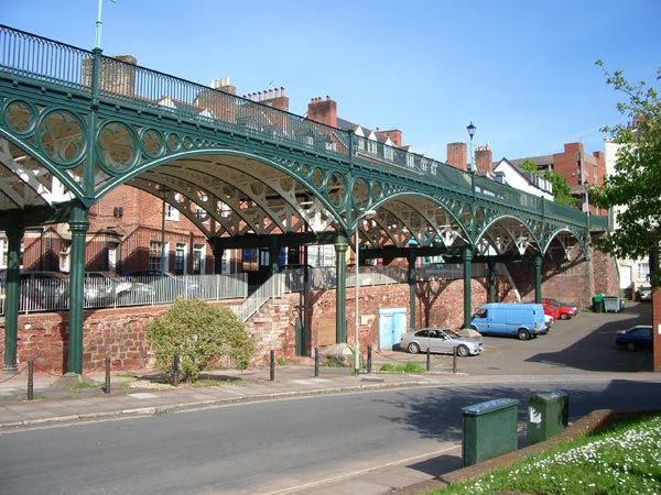 Photograph taken with the Pentax Optio S5n camera featuring a green ornate metal bridge with intricate designs over a road, with red-bricked buildings and parked cars in the background on a sunny day.