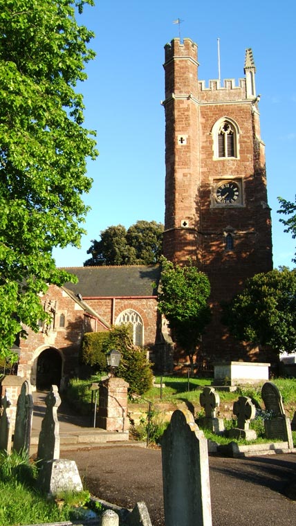 Photograph of an old stone church with a tall tower and clock, surrounded by a graveyard with headstones, captured in bright sunlight, taken with a Fujifilm FinePix F810 Zoom camera.