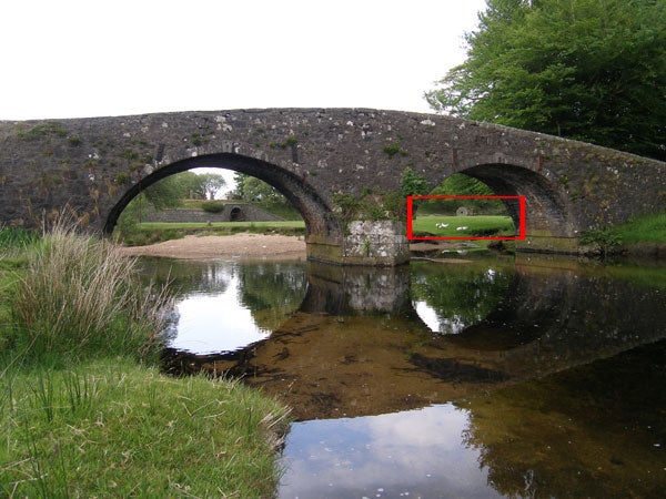 A serene scene captured by the Olympus Camedia C-7070, featuring an old stone bridge with double arches reflected in the calm river below, surrounded by lush greenery, with a red box highlighting the detailed area in the photo.