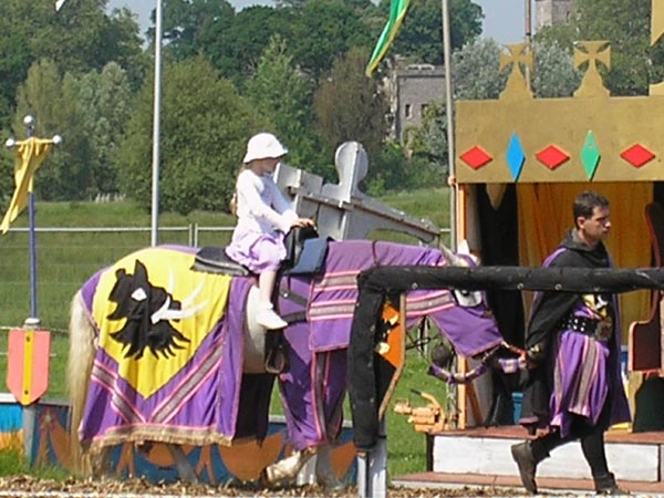 A child wearing a safety helmet sitting on a fake horse at a medieval-themed event, with a figure in knight armor nearby and colorful tents in the background, captured with the Olympus C-180 camera.