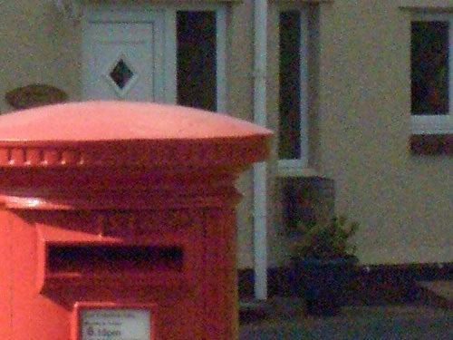 Close-up photo of an orange postal box with a blurred building in the background, possibly taken using the Casio EX-P505 to illustrate camera quality.