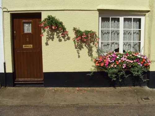 Photograph taken with a Casio EX-P505 camera showing a quaint exterior of a house with a wooden front door flanked by windows, adorned with pink and red flowers growing on the walls and a vibrant flower box.