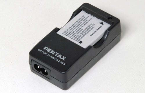 Battery charger for Pentax Optio S5z camera with a label showing, placed on a light background.
