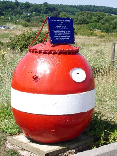 A large red spherical buoy with a white horizontal stripe, placed on land with a commemorative plaque attached to it, with a natural landscape in the background.