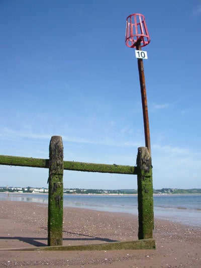 A clear image of a beachscape taken with the Pentax Optio S5z, featuring a rusty metal structure with green algae, a red basket at the top, and a sign with the number 10, under a blue sky.