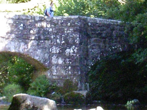 A blurry photograph of an old stone bridge over a stream with a person sitting on the edge, possibly taken with a low-resolution setting on the Pentax Optio SVi digital camera.