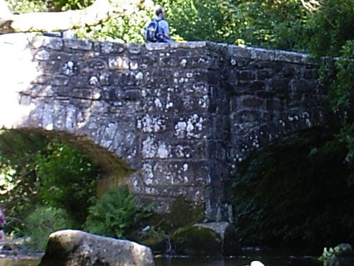 Photo taken with Pentax Optio SVi digital camera featuring an old stone bridge over a stream with a person sitting on top of the bridge, surrounded by lush greenery. The image showcases the camera's outdoor photography capability, with a focus on the texture of the bridge and the natural environment.