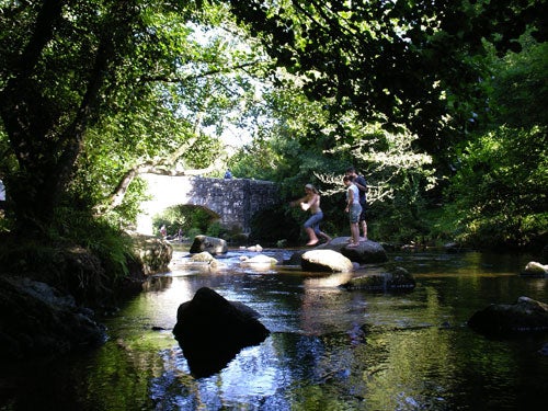 Photo taken with Pentax Optio SVi digital camera featuring an old stone bridge over a stream with a person sitting on top of the bridge, surrounded by lush greenery. The image showcases the camera's outdoor photography capability, with a focus on the texture of the bridge and the natural environment.Photo captured by the Pentax Optio SVi digital camera showcasing two individuals stepping on stones across a serene river with a backdrop of lush foliage and a stone bridge.