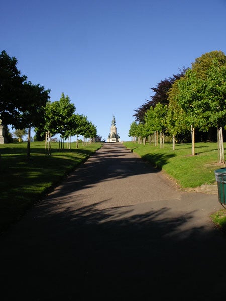 Photograph taken with a Pentax Optio SVi digital camera showing a tree-lined pathway leading to a statue with clear blue skies above.