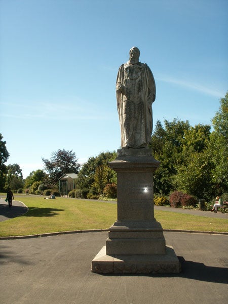 Stone statue of a historical figure on a pedestal in a park with trees and a clear blue sky.