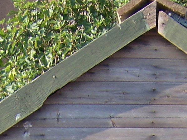 Close-up of a wooden structure with a triangular section, possibly part of a fence or decking, with green shrubbery in the background, with the image demonstrating the picture quality of the Kodak Easyshare V550 camera.