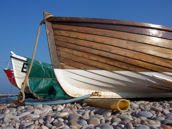 A wooden rowboat with a rope tied to the bow, sitting on a pebble beach with a blue sky in the background, possibly taken with a Kodak Easyshare V550 camera.