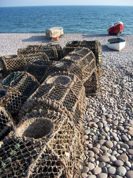Traditional lobster traps resting on a pebble beach with two small boats in the background by the sea.