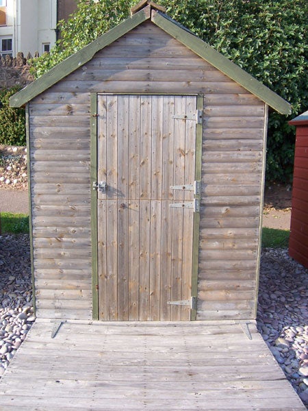 Wooden garden shed with closed door and ramp leading up to entrance, surrounded by gravel and a hedge in the background.