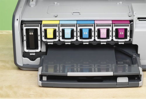 HP Photosmart 8250 printer with open cover showing six individual ink cartridges in a color-coded arrangement and an empty paper tray in the foreground.