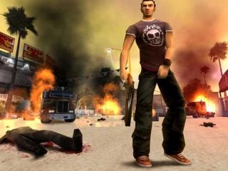 In-game screenshot from Total Overdose showing the main character standing in the center with a gun in his right hand. Behind him, there are explosions, a burning vehicle, and a downed enemy on the ground amidst a street scene with palm trees and various storefronts.