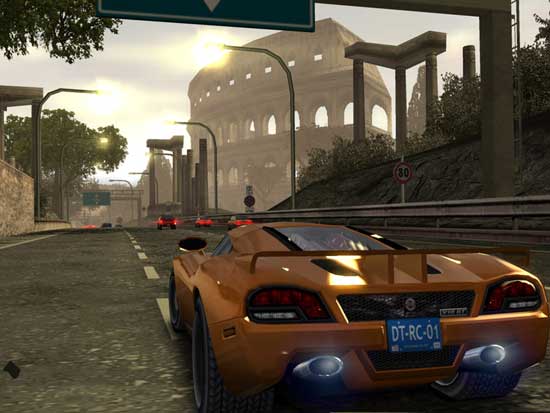 Screenshot from the video game Burnout: Revenge featuring a rear view of a playable orange sports car racing through a city street course with other vehicles in the distance and a large Colosseum-like structure in the background.