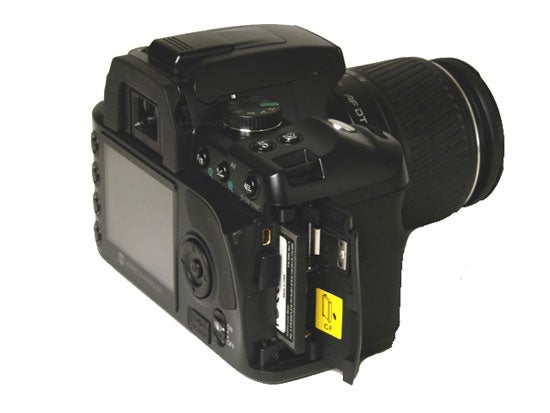 Konica Minolta Dynax 5D digital SLR camera with a black lens, shown from the back with memory card slots open and displaying a compact flash card inserted.