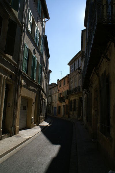 Photo taken with a Konica Minolta Dynax 5D digital SLR camera, featuring a sunlit European-style narrow street with tall, shadowed buildings and balconies, demonstrating the camera's dynamic range.