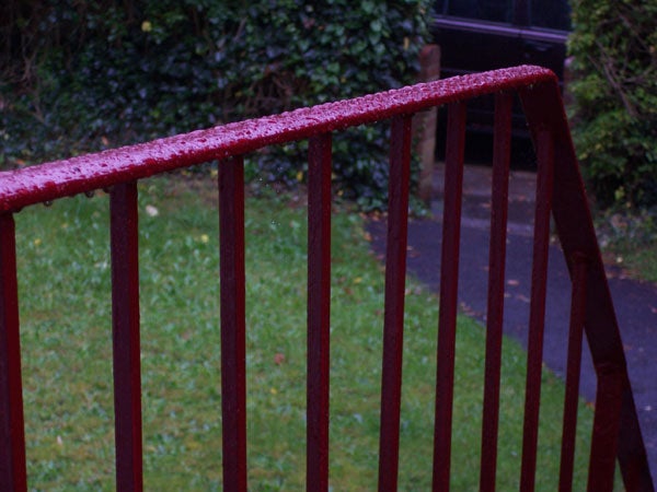 Red metal railing with dewdrops on top, with a blurred green garden in the background, potentially demonstrating the depth-of-field capability of the Kodak EasyShare C360 camera.