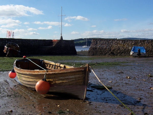 A wooden rowboat with orange buoys tied to it, resting on a muddy shore with a low stone wall and docked boats in the background under a partly cloudy sky.