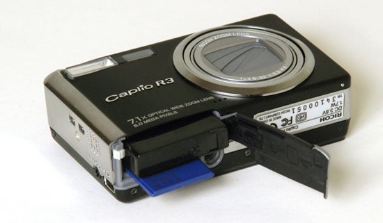 A Ricoh Caplio R3 digital camera with the battery compartment open showing the blue battery inside.