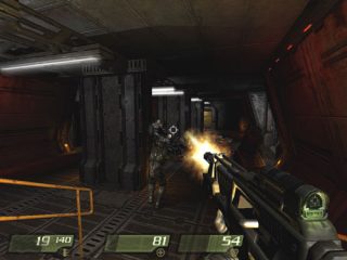 First-person perspective screenshot from the video game Quake 4 showing the player character firing a weapon at an enemy soldier within a dimly lit, industrial-style corridor.