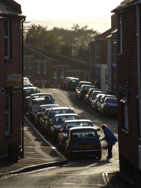 A street view captured in early morning light, showing parked cars lining both sides of the road with a person inspecting a car, hinting at the dynamic range and zoom capabilities of the Fujifilm FinePix S9500 camera.