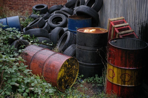 Photograph showing a cluttered scene with multiple old tires stacked in the background, rusted barrels in the foreground, and overgrown vegetation, showcasing the image quality and color reproduction of the Epson R-D1 Digital Rangefinder Camera.