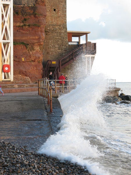 A wave crashes against the seawall by a red rock formation with a lookout platform while pedestrians observe from a safe distance.