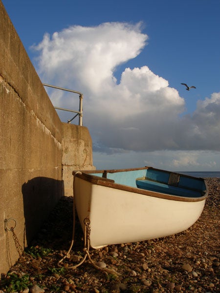 White and blue rowboat moored on a pebble beach next to a concrete wall with a metal railing, under a blue sky with scattered clouds and a flying seagull.