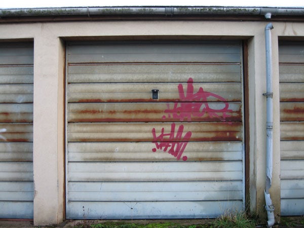 The image shows a weathered garage door with streaks of rust and pink graffiti visible on the surface. There is a small black square that could potentially be a camera, perhaps the Canon Digital IXUS 750, mounted on the door's exterior.