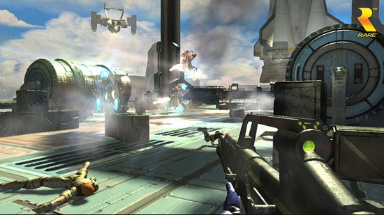 A screenshot from the video game Perfect Dark Zero showing a first-person perspective with a gun aimed at an enemy character who is laying on the ground in a futuristic setting with bright lighting, industrial structures, and flying vehicles in the background. The game's developer logo, Rare, is visible in the corner.