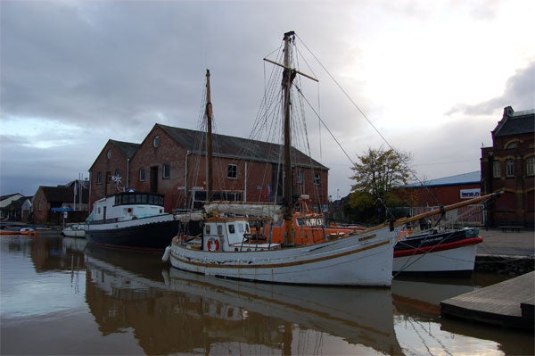 A Nikon D50 + 18-55mm Lens captured image depicting an overcast sky with subdued lighting, featuring an old sailing ship docked at a calm waterway in front of traditional brick buildings, reflecting a serene maritime scene.