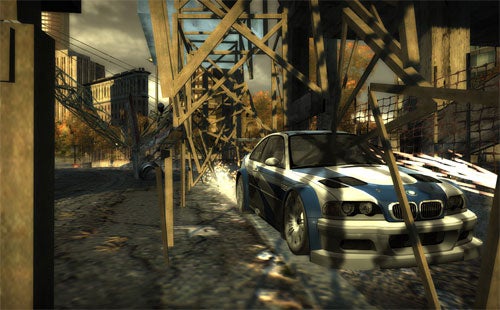 A screenshot from the video game Need for Speed: Most Wanted 2005 showing a BMW M3 GTR racing through an industrial area with a construction scaffold and autumn-colored trees in the background.