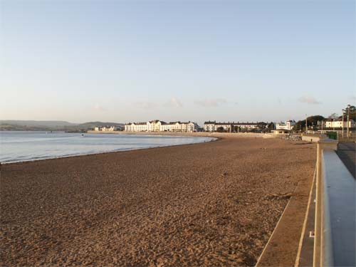 Photo captured with Konica Minolta Dimage Z6 showing a scenic beach with a clear sky, the sea on the left, and a line of buildings along the coastline in the distance.
