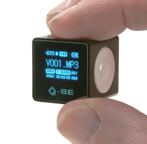 A close-up of a person holding a small, cube-shaped SupportPlus Q-BE MP3 player with a digital screen displaying the track information.