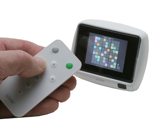 A person holding a white iRiver U10 digital media player with its screen displaying a colorful grid menu, next to a matching white docking station.