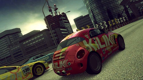 Screenshot from Ridge Racer 6 video game showing two vibrant racing cars with decals competing on an urban track with buildings in the background and construction cranes in the skyline.