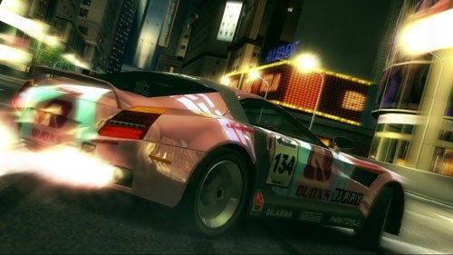 A screenshot from the racing video game Ridge Racer 6, showing a pink and white race car with the number 134 and sponsorship decals speeding through a city at night, with motion blur effects highlighting its speed.