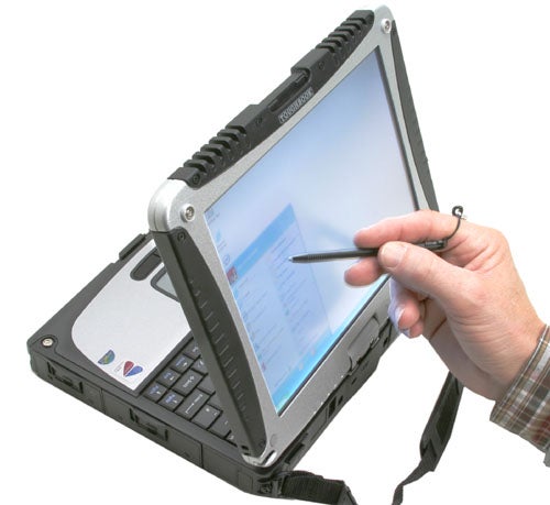 A Panasonic ToughBook CF-18 rugged notebook in tablet mode with a person using a stylus on the touchscreen display. The device has a black protective case and visible ports, with a hand strap on the back for easy handling.