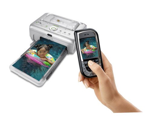 A Kodak EasyShare Printer Dock Plus Series 3 with a printed photo emerging, beside a hand holding a phone displaying the same image.