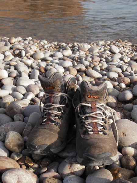 A pair of hiking boots on a pebble-covered river shore, suggesting an outdoor scene potentially captured with a Pentax Optio WPi - Waterproof Camera due to the camera's suitability for nature and adventure photography.