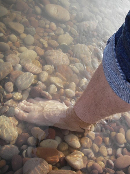 Person's lower leg and foot submerged in clear water above a riverbed with smooth stones, demonstrating the underwater clarity achievable with the Pentax Optio WPi waterproof camera.