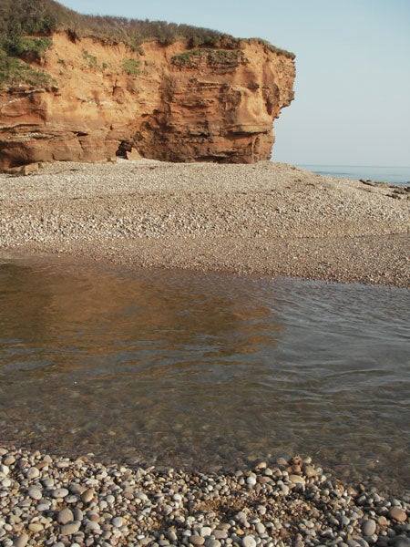 Photo taken with Pentax Optio WPi showing a clear image of a rocky beach with pebbles in the foreground, calm water in the middle, and a red clay cliff in the background against a clear sky.